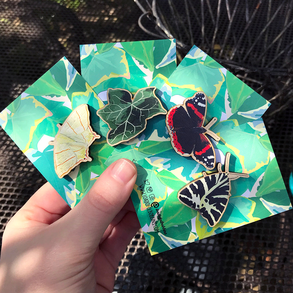 A collection of laser cut wooden pins showing illustration by Loadofolbobbins of a swallowtail moth, a dark green ivy leaf, a red admiral butterfly and a jersey tiger moth. The pins are mounted on a backing card with a repeat ivy leaf pattern.