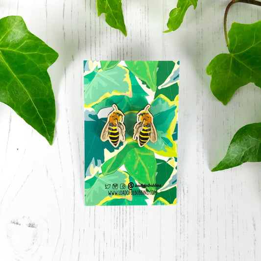 A pair of laser cut matching wooden stud earrings showing an illustration by Loadofolbobbins of an ivy mining bee. The earrings are mounted on a backing card with a repeat ivy leaf pattern.