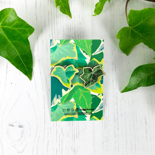 A pair of laser cut mix and match wooden stud earrings showing illustrations by Loadofolbobbins of a light green ivy leaf and a dark green ivy leaf. The earrings are mounted on a backing card with a repeat ivy leaf pattern.