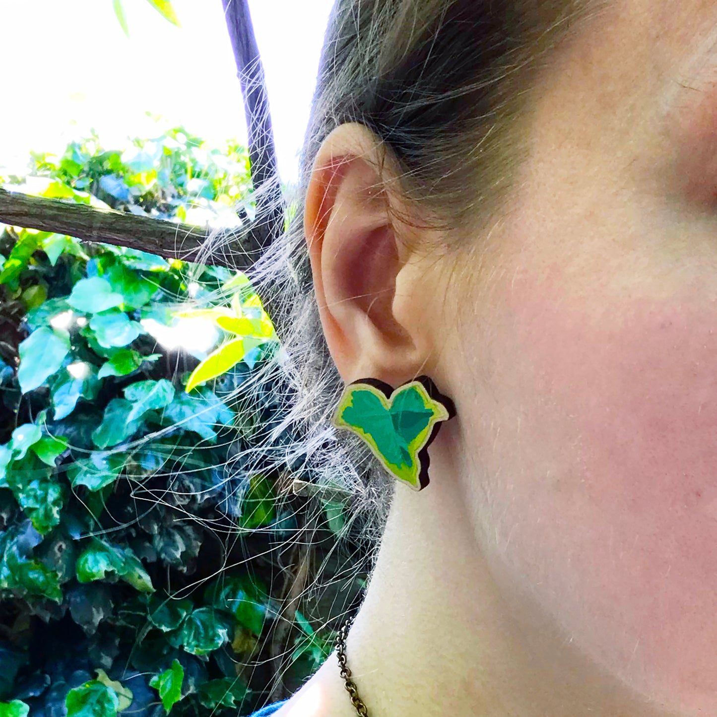 A close up photo of a white woman's ear with blondish brown hair worn up and standing against a wall of ivy. The main focus of the image is her ear which is wearing a small laser cut wooden stud earring of a light green ivy leaf by Loadofolbobbins.