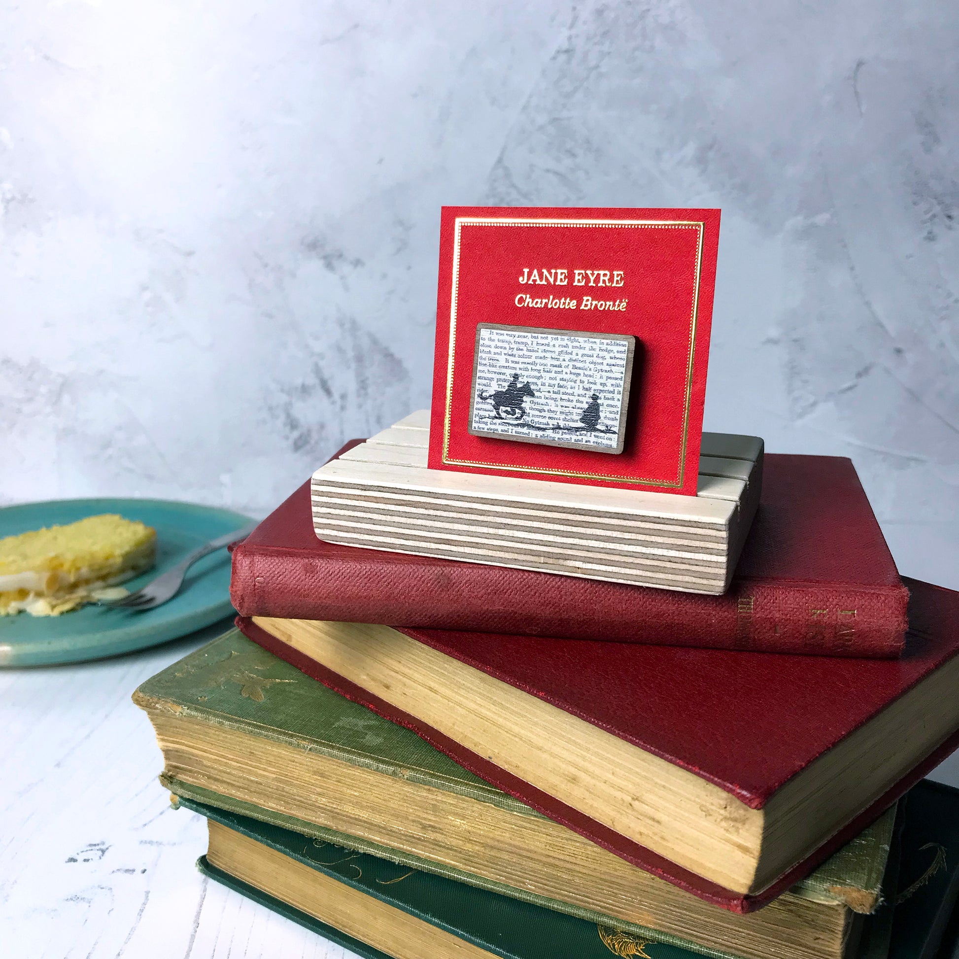 A laser cut wooden brooch by loadofolbobbins showing a silhouette scene inspired by the novel Jane Eyre by Charlotte Brontë. The scene shows a silhouette scene of a man galloping on a horse towards a woman, Mr Rochester and Jane. They are in Victorian dress. The scene is printed against original text from the novel. The necklace is mounted on a red backing card with gold foil lettering detail and made to look like the front of a book.