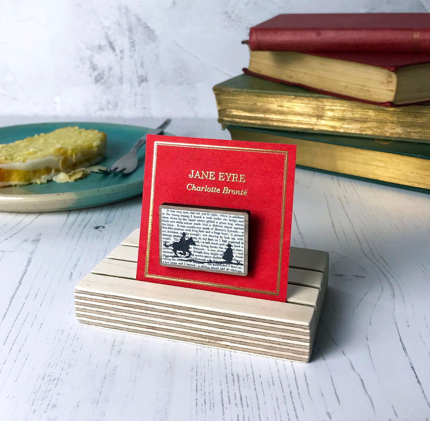 A laser cut wooden brooch by loadofolbobbins showing a silhouette scene inspired by the novel Jane Eyre by Charlotte Brontë. The scene shows a silhouette scene of a man galloping on a horse towards a woman, Mr Rochester and Jane. They are in Victorian dress. The scene is printed against original text from the novel. The necklace is mounted on a red backing card with gold foil lettering detail and made to look like the front of a book.