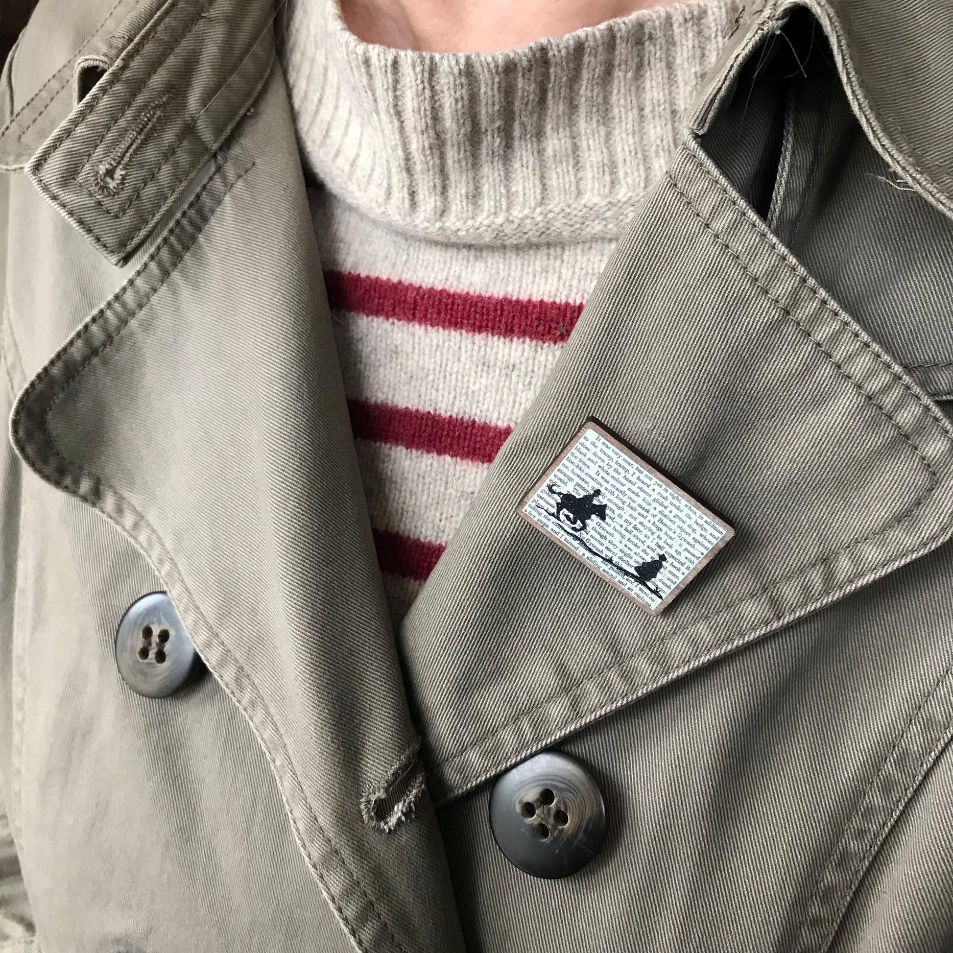 A laser cut wooden brooch by loadofolbobbins showing a silhouette scene inspired by the novel Jane Eyre by Charlotte Brontë. The scene shows a silhouette scene of a man galloping on a horse towards a woman, Mr Rochester and Jane. They are in Victorian dress. The scene is printed against original text from the novel. The brooch is being worn on the lapel of a jacket.