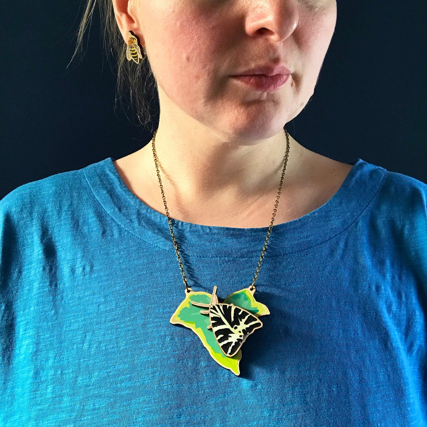 Jersey Tiger Moth and Ivy  necklace by Loadofolbobbins. A laser cut wooden necklace that shows a ivy leaf with a Jersey Tiger moth on. The necklace hangs on an antique bronze chain. It is being worn by me a white woman wearing a light blue top. You can just see the bottom of her face and neck.