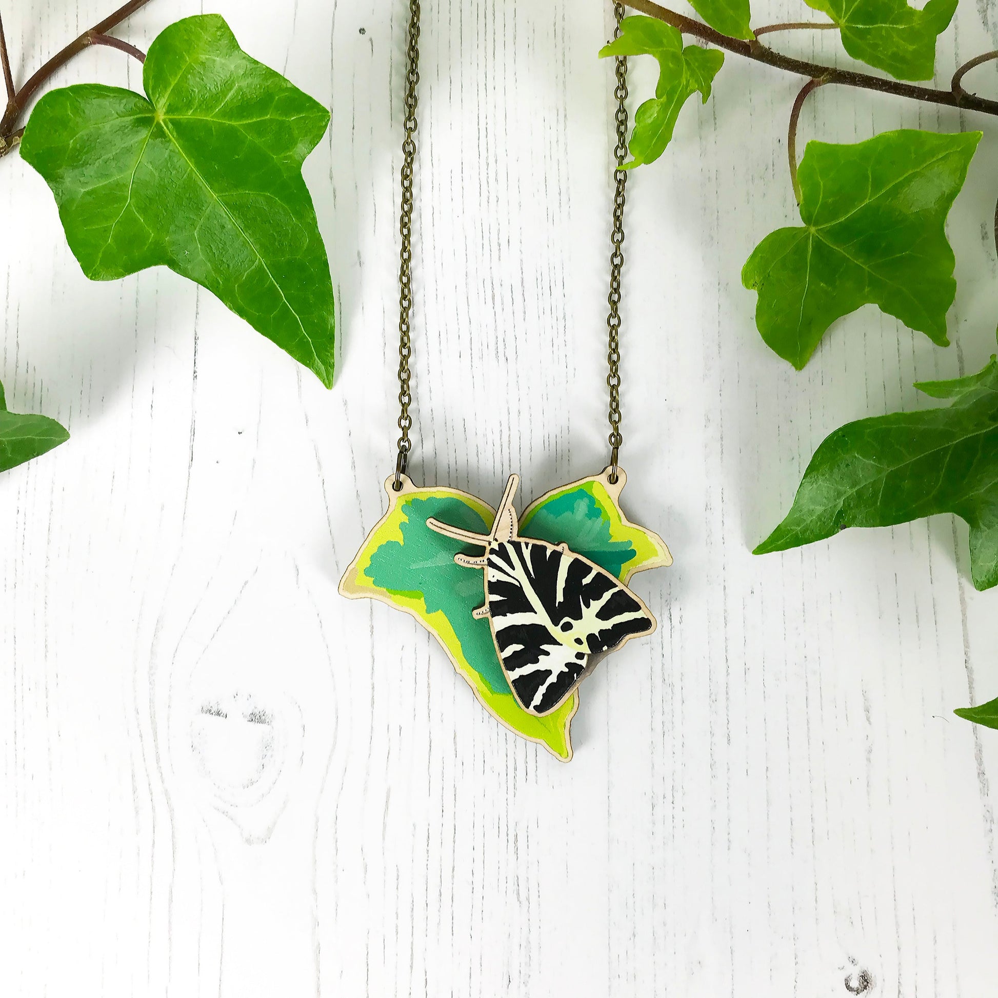 A laser cut wooden statement necklace showing an illustration of an variegated ivy leaf and Jersey Tiger Moth by loadofolbobbins. The moth stands proud of the necklace in a 3D relief effect.  The wooden necklace is hanging from an antique bronze cable chain.