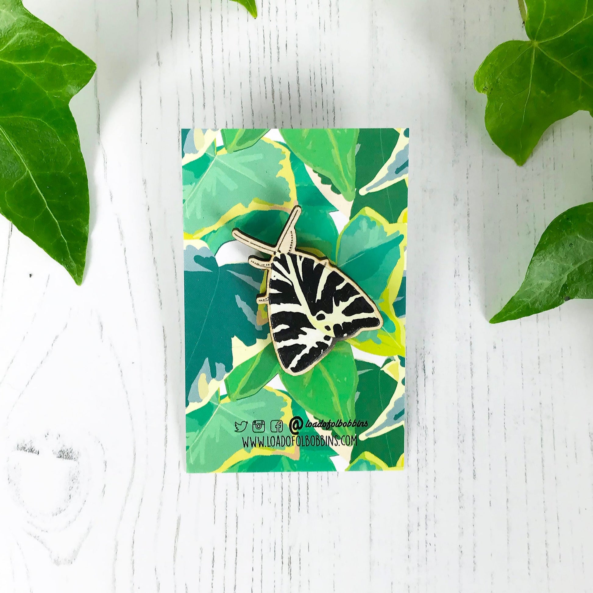A laser cut wooden brooch showing an illustration by Loadofolbobbins of a Jersey Tiger moth. The brooch is mounted on a backing card with a repeat ivy leaf pattern.