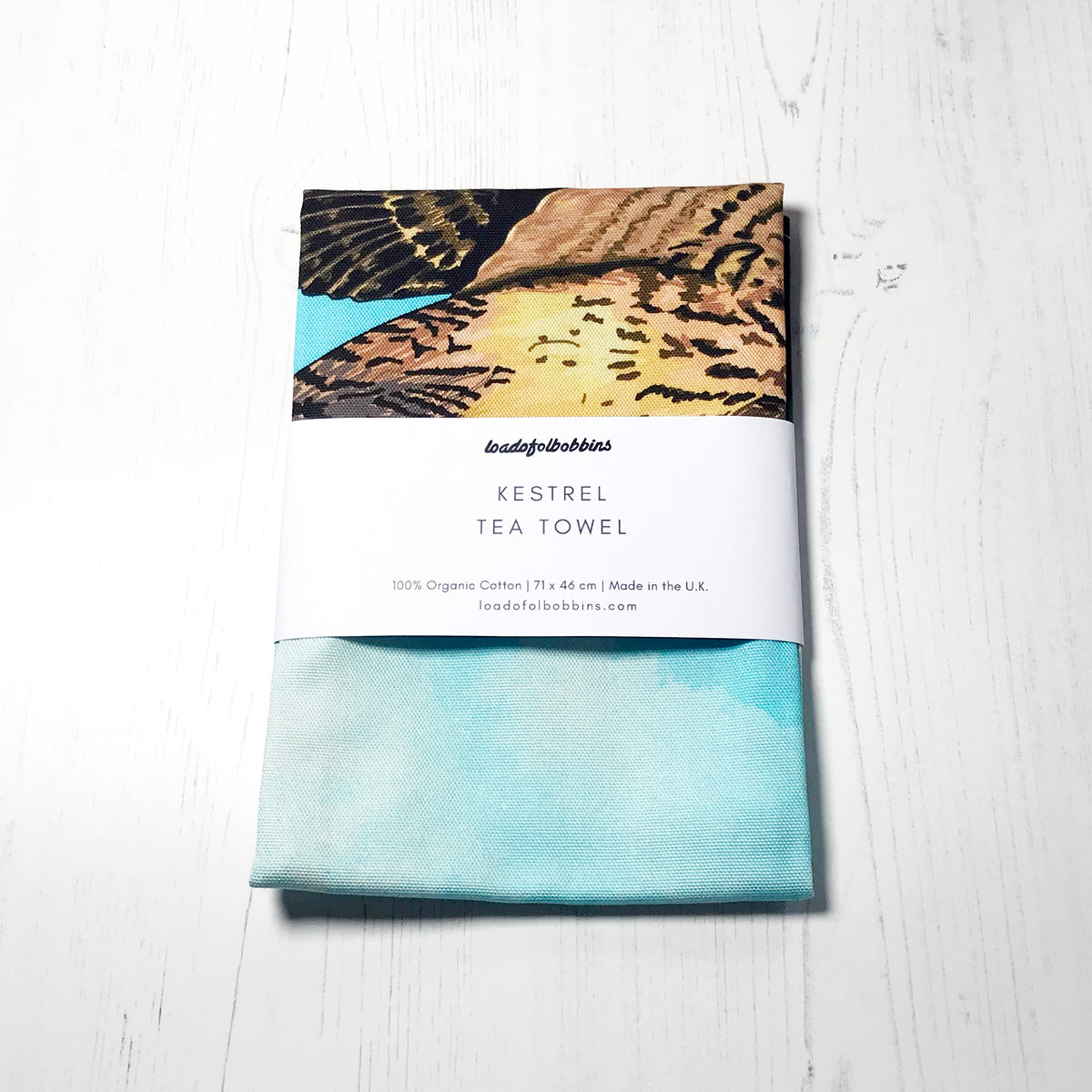A folded bird themed illustration tea towel by loadofolbobbins. It shows a hovering kestrel flying above tree tops against an inky wash turquoise and white background.