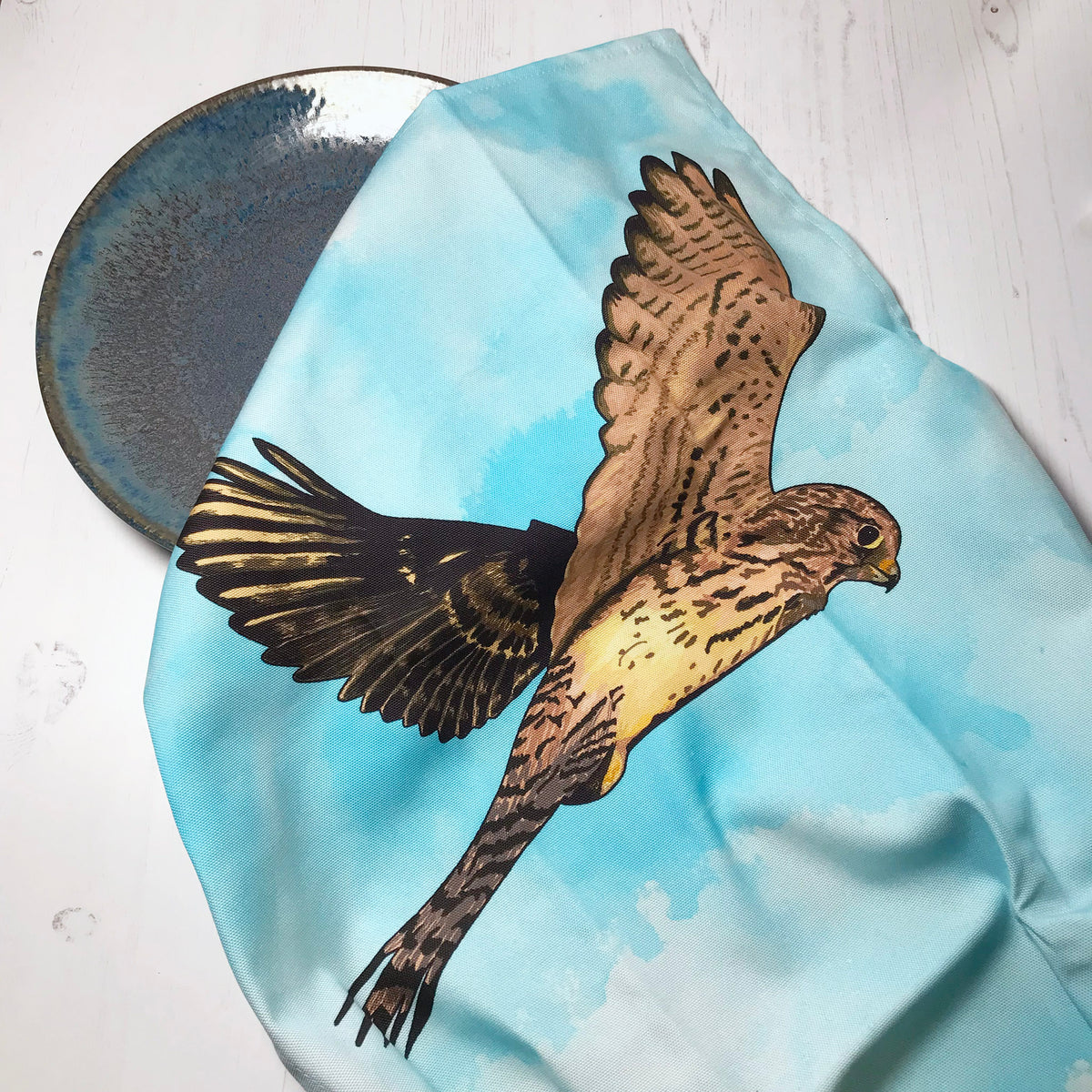 A  photo of a loosely scrunched up tea towel looking like it is drying a ceramic glazed dark turquoise plate. The tea towel shows an illustration of a hovering kestrel flying above the tops of trees,  against an inky wash turquoise and white background.