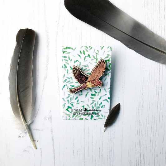 A laser cut wooden brooch showing an illustration by Loadofolbobbins of a flying kestrel with wings spread out wide, hovering. The brooch is mounted on a backing card with a repeat abstract watercolour leaf pattern.