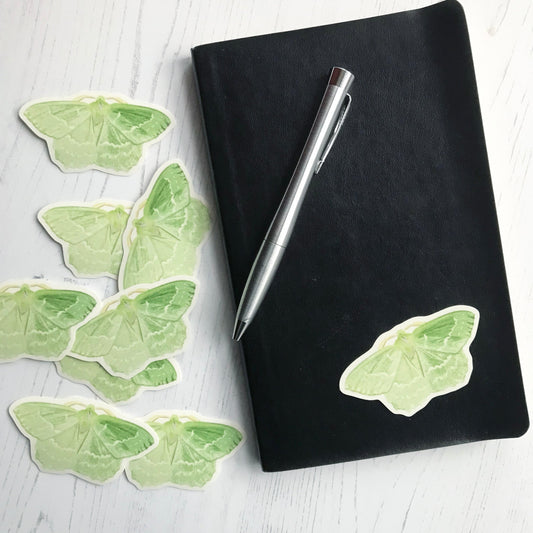 A large emerald moth sticker created from an original digital illustration by Loadofolbobbins. The moth is a beautiful light green allover wings and fluffy body and head. On both sets of light green wings there are a series of subtle cream markings in a wavy striped pattern towards the bottom of the wings. It's fairly thick feathered antennae are a light yellow in colour. The sticker is on a black notebook with a pen resting on it.