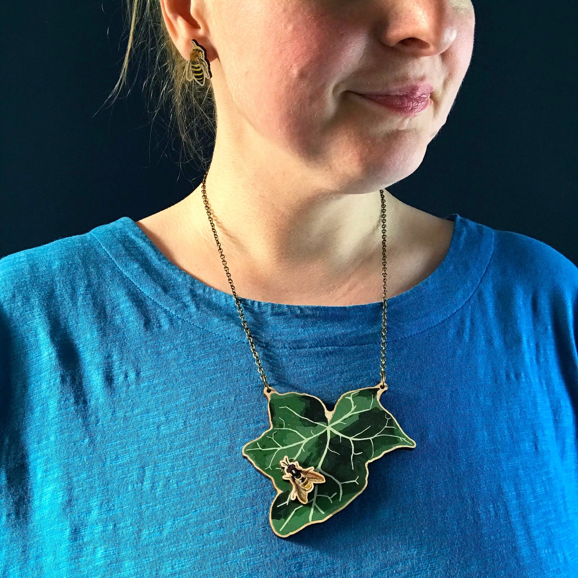 Golden Hoverfly and Ivy  necklace by Loadofolbobbins. A laser cut wooden necklace that shows a large ivy leaf with a golden hoverfly on. The necklace hangs on an antique bronze chain. It is being worn by me a white woman wearing a light blue top. You can just see the bottom of my face and neck.