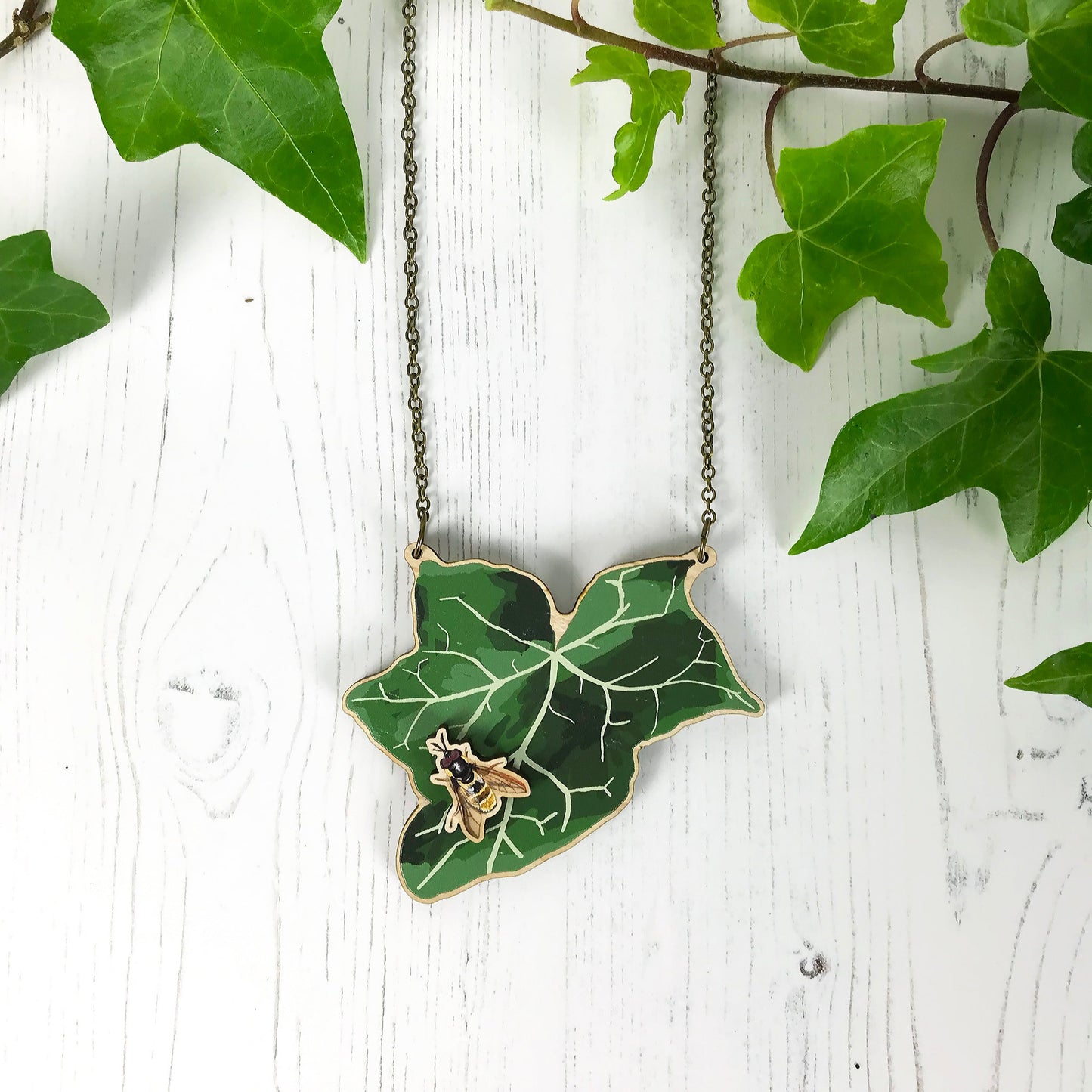 A laser cut wooden statement necklace showing an illustration of an dark green ivy leaf and golden hoverfly by loadofolbobbins. The hoverfly stands proud of the necklace in a 3D relief effect.  The wooden necklace is hanging from an antique bronze cable chain.