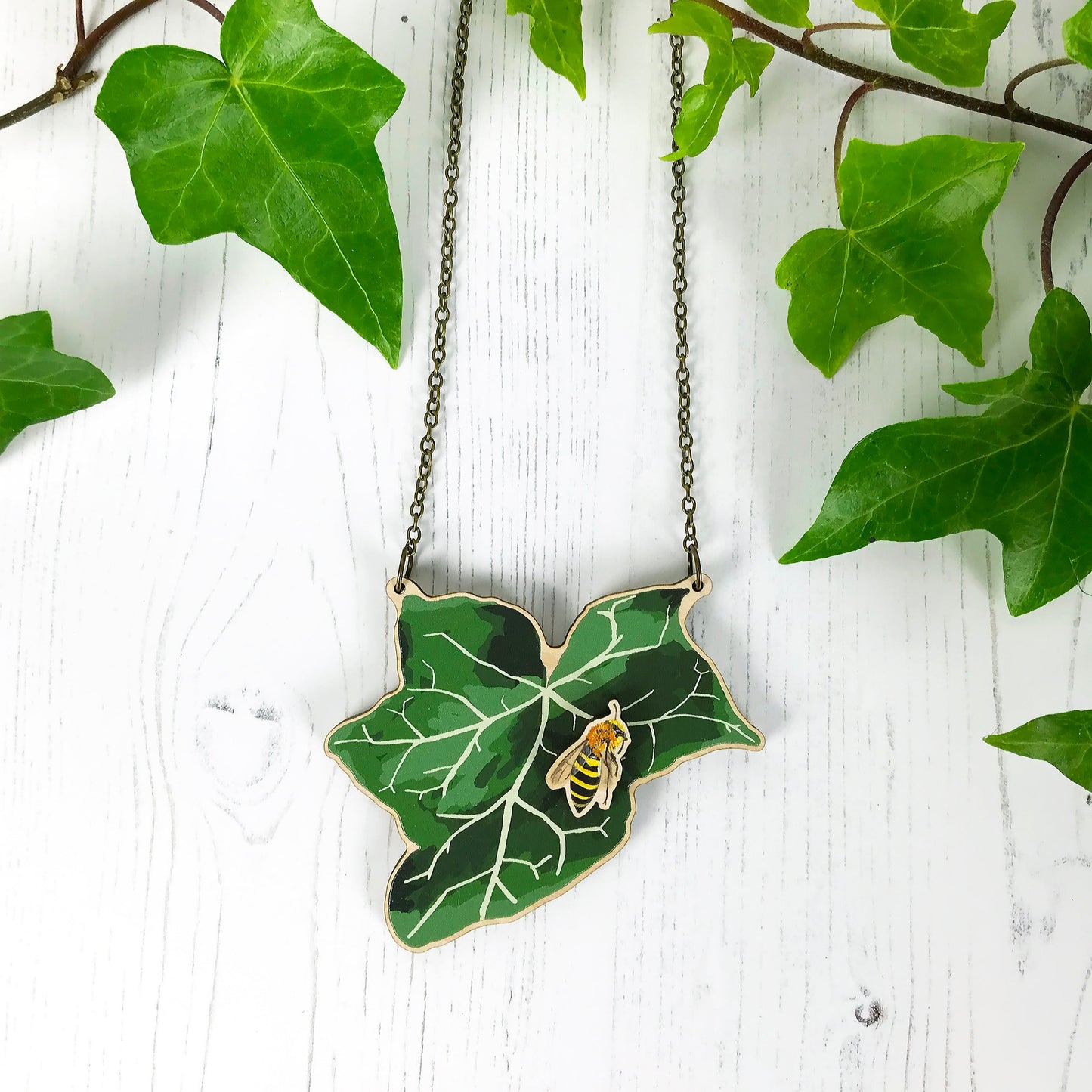 A laser cut wooden statement necklace showing an illustration of an dark green ivy leaf and an ivy mining bee by loadofolbobbins. The bee stands proud of the necklace in a 3D relief effect.  The wooden necklace is hanging from an antique bronze cable chain.
