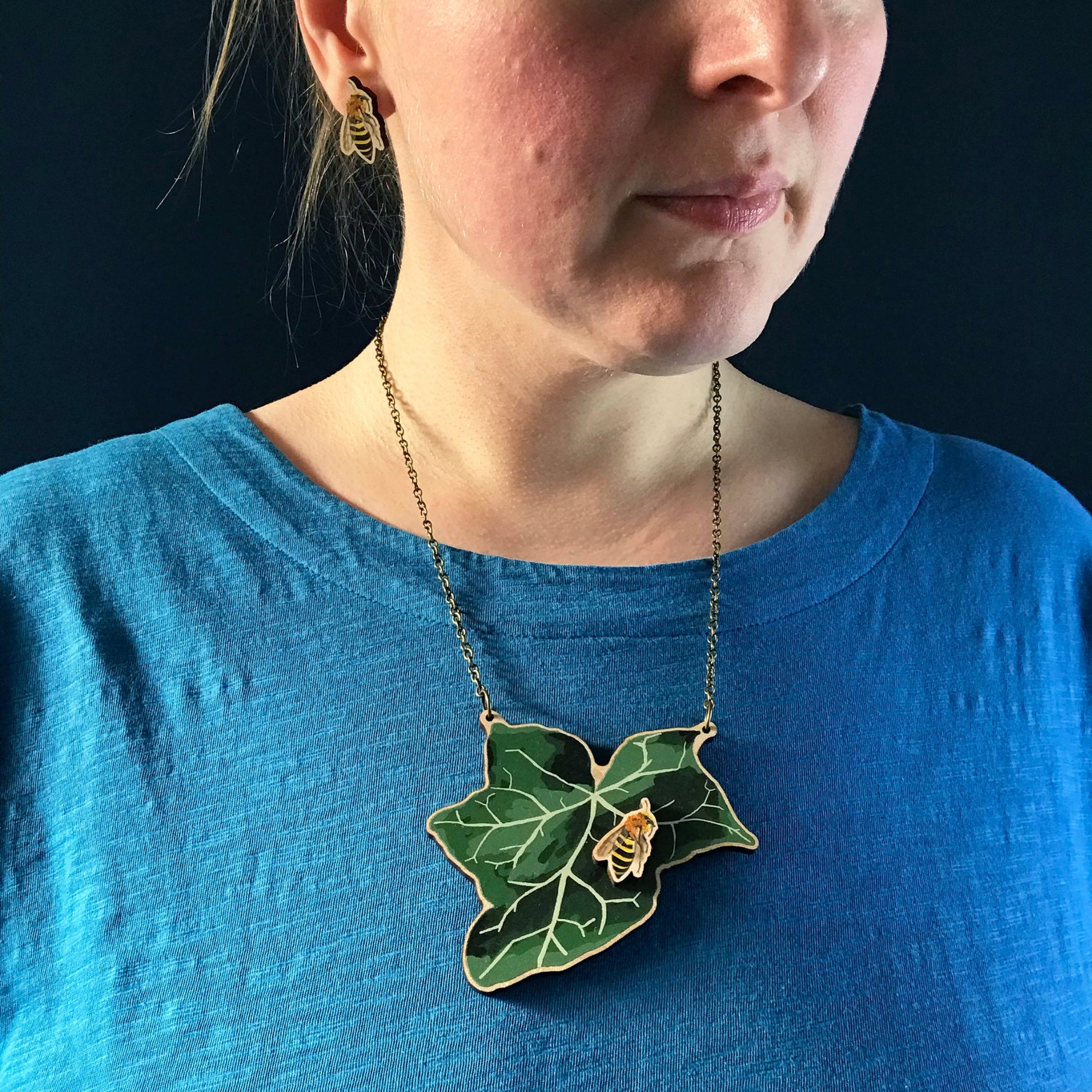 Ivy Mining Bee and Ivy necklace by Loadofolbobbins. A laser cut wooden necklace that shows a large ivy leaf with a ivy mining bee on. The necklace hangs on an antique bronze chain. It is being worn by me a white woman wearing a light blue top. You can just see the bottom of my face and neck.