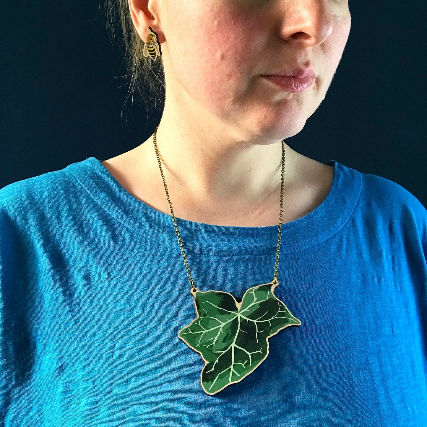A laser cut wooden statement necklace showing an illustration of an dark green ivy leaf by loadofolbobbins. The wooden necklace is hanging from an antique bronze cable chain and worn against a blue top.