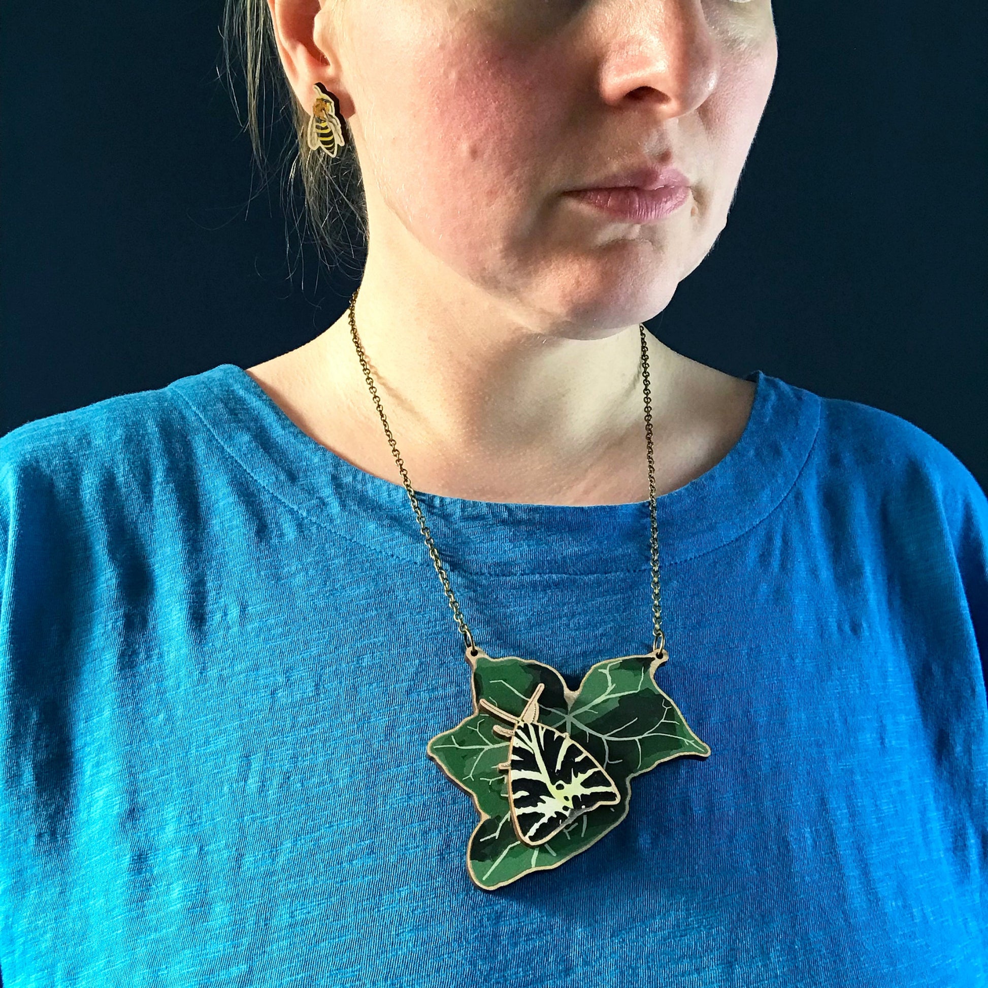 Jersey Tiger Moth and Ivy  necklace by Loadofolbobbins. A laser cut wooden necklace that shows a large ivy leaf with a swallowtail moth on. The necklace hangs on an antique bronze chain. It is being worn by me a white woman wearing a light blue top. You can just see the bottom of my face and neck.