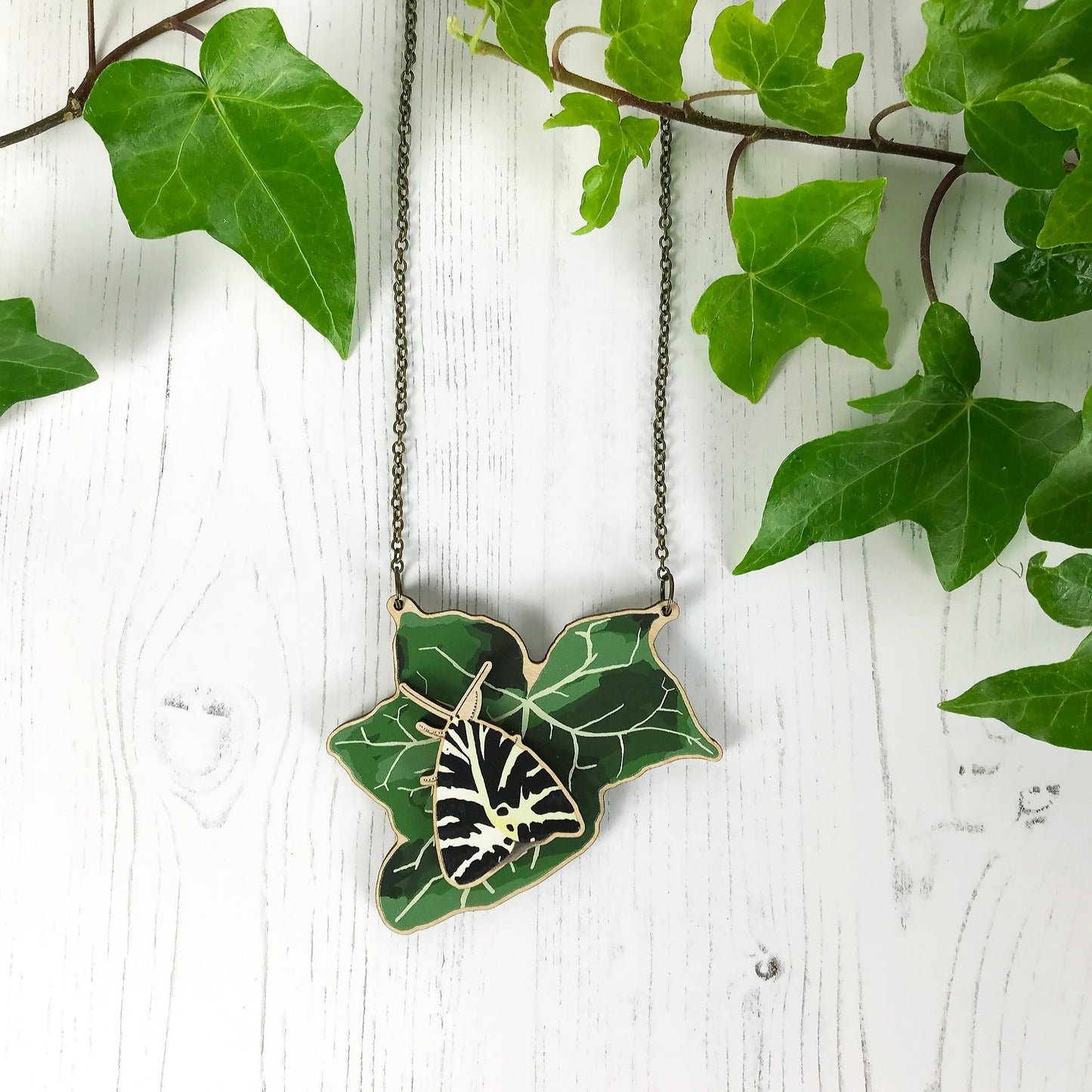 A laser cut wooden statement necklace showing an illustration of an dark green ivy leaf and Jersey Tiger Moth by loadofolbobbins. The moth stands proud of the necklace in a 3D relief effect.  The wooden necklace is hanging from an antique bronze cable chain.