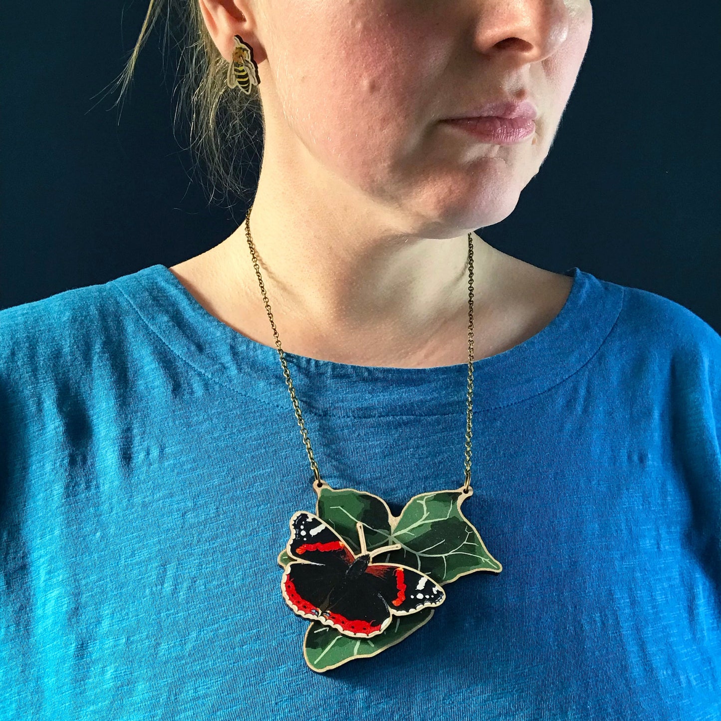 Red Admiral Butterfly and Ivy  necklace by Loadofolbobbins. A laser cut wooden necklace that shows a large ivy leaf with a red admiral butterfly on. The necklace hangs on an antique bronze chain. It is being worn by me a white woman wearing a light blue top. You can just see the bottom of my face and neck.