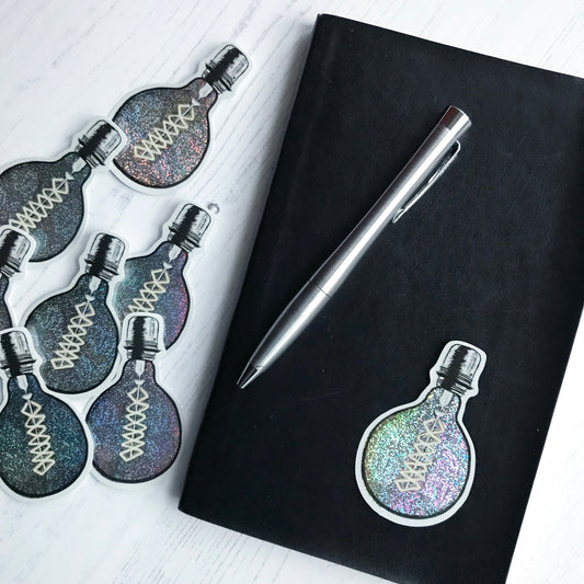 A sticker by Loadofolbobbins of a light bulb with a bold black outline design, glowing filament and silver holographic glitter effect on the main bulb.