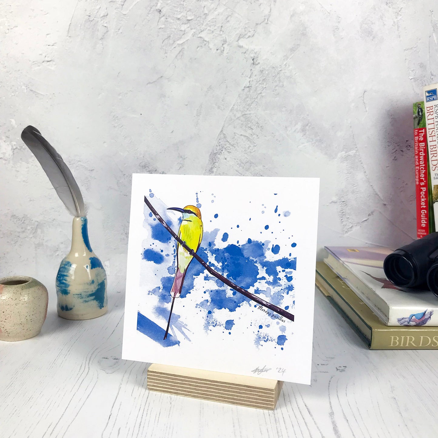 A photo of a small sized 14 cm square art print made from an original illustration by Loadofolbobbins. It is of a little bee eater bird perched on a fine curved branch. They have vibrant green upper feathers and yellow head. there's a black band of feathers like a zorro mask. It has a long pointed curved grey beak. The background of the image is made of rich bright blue inky splash marks. There is a 2.5 cm white border for framing.