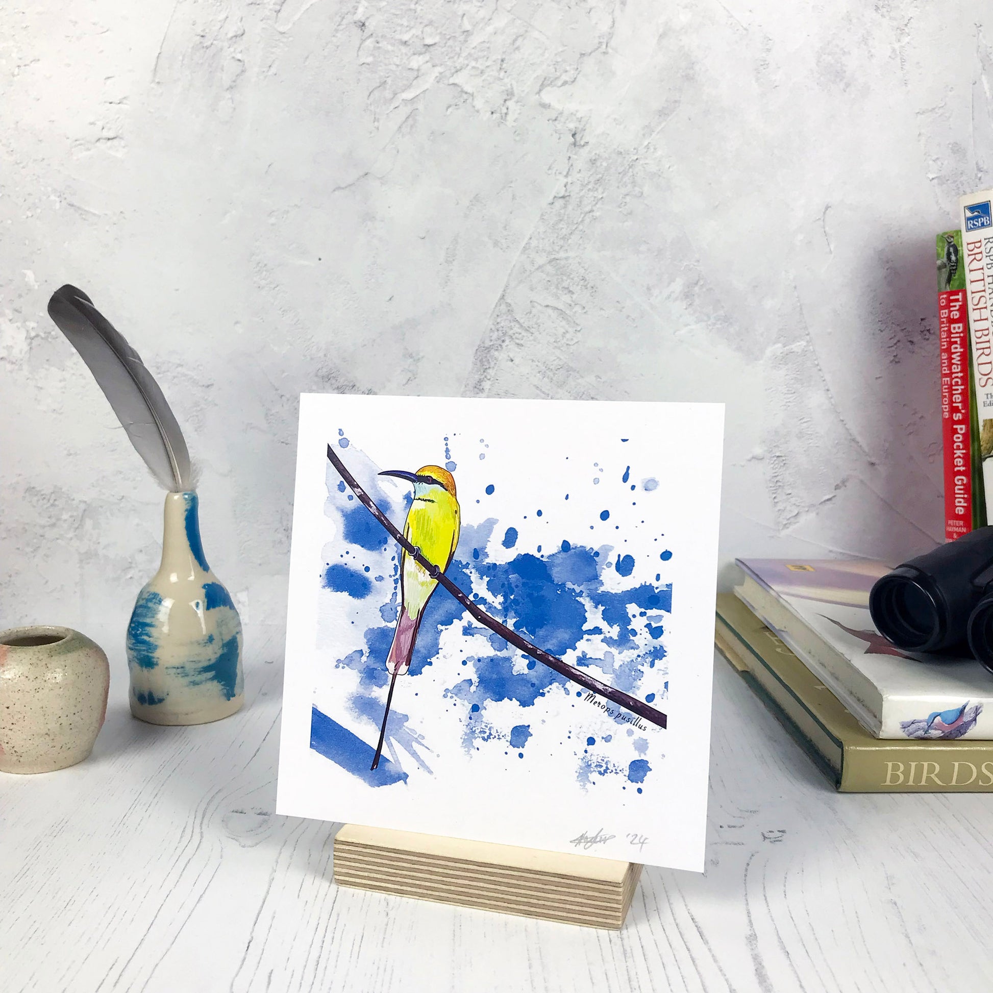 A photo of a small sized 14 cm square art print made from an original illustration by Loadofolbobbins. It is of a little bee eater bird perched on a fine curved branch. They have vibrant green upper feathers and yellow head. there's a black band of feathers like a zorro mask. It has a long pointed curved grey beak. The background of the image is made of rich bright blue inky splash marks. There is a 2.5 cm white border for framing.