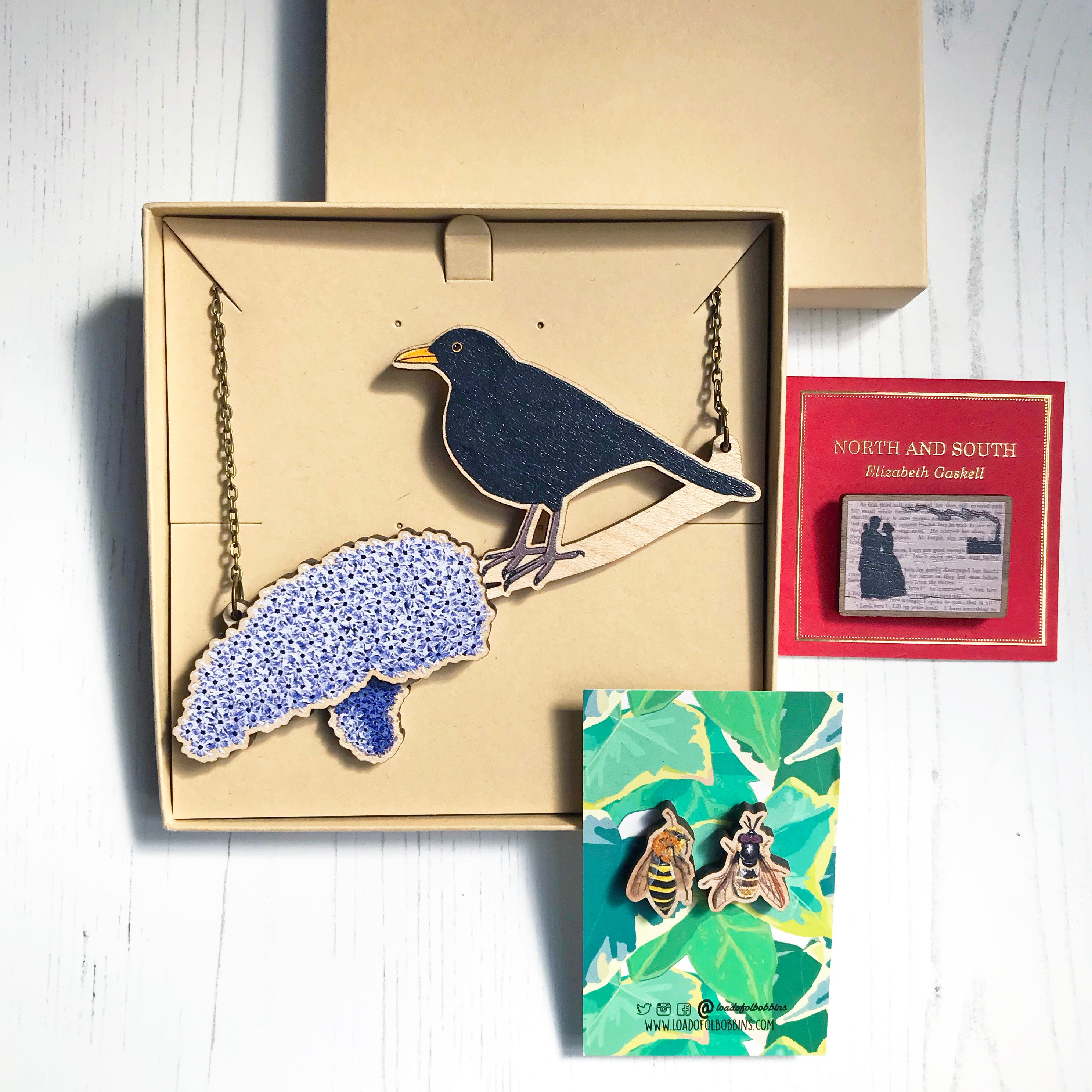 A photo of a selection of products available from Loadofolbobbins highlighting the quality and lovely packaging.  Top left is a wooden necklace showing a male blackbird perching on a branch of lilac blossom, it's packaged in a lovely presentation card board box. To the right of this is a North and South classic literature brooch which is attached to a gold foiled red backing card. At the bottom of the image is a mix and match pair of studs showing a bee and hoverfly mounted of colourful backing card.