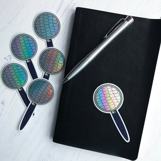 A holographic effect magnifying glass sticker by Loadofolbobbins. The sticker is of a magnifying glass with a blue handle and withing the glass of it there is close up black outline drawings of moth wing scales. The background of the scales drawing is holographic.