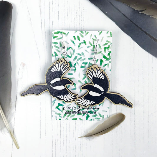 A pair of laser cut wooden dangle earrings showing an illustration by Loadofolbobbins of a flying magpie. The earrings are mounted on a backing card with a repeat abstract watercolour leaf pattern.