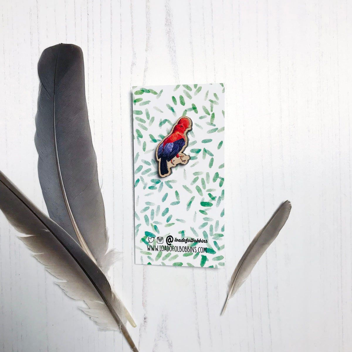 A 30mm mini wooden lapel pin of a Andean cock-of-the-rock bird perched on a branch and  mounted on an illustrated backing card. The pin is made from an original illustration and is detailed and expressive in style. The bird is orange and black with a distinct circular crest on it's head. The backing card has a repeat green watercolour effect pattern that looks like leaves.
