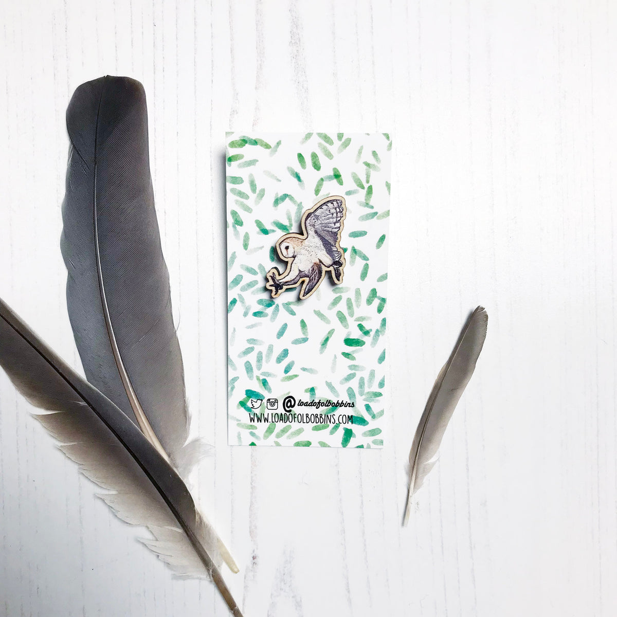 A 30mm mini wooden lapel pin of a swooping barn owl mounted on an illustrated backing card. The pin is made from an original illustration and is detailed and expressive in style. The bird is creams and browns with wings in full flight. The backing card has a repeat green watercolour effect pattern that looks like leaves.