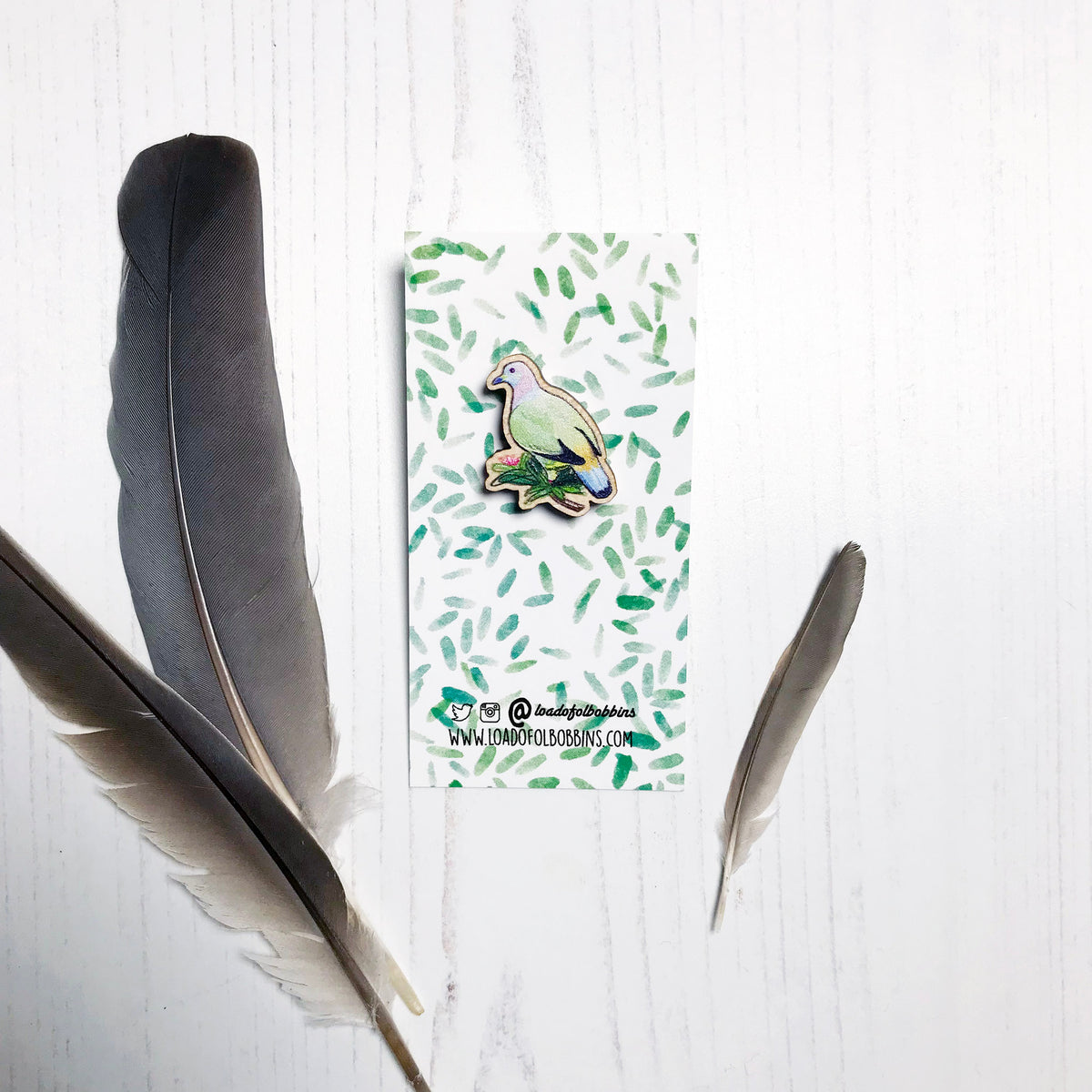 A 27mm mini wooden lapel pin of a pink-necked green pigeon mounted on an illustrated backing card. The pin is made from an original illustration and is detailed and expressive in style. The bird pastel markings in greens, pinks, yellows and blues and is perched on a lush green branch. The backing card has a repeat green watercolour effect pattern that looks like leaves.