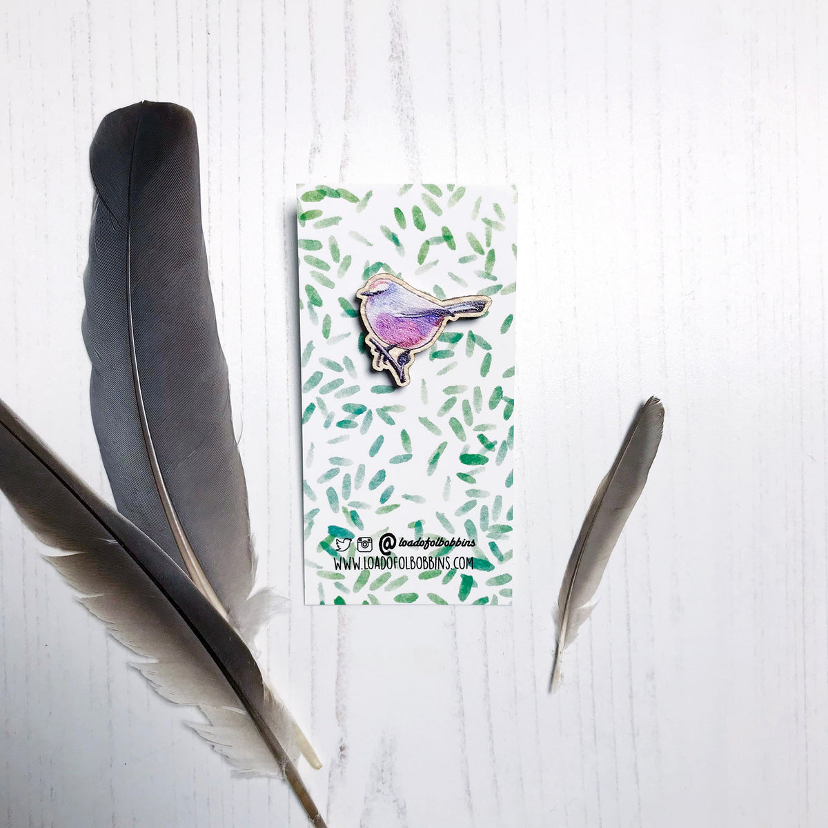 A 26mm mini wooden lapel pin of a white-browed tit warbler mounted on an illustrated backing card. The pin is made from an original illustration and is detailed and expressive in style. The bird is a mix of amazing pinks, purples and blues. The backing card has a repeat green watercolour effect pattern that looks like leaves.