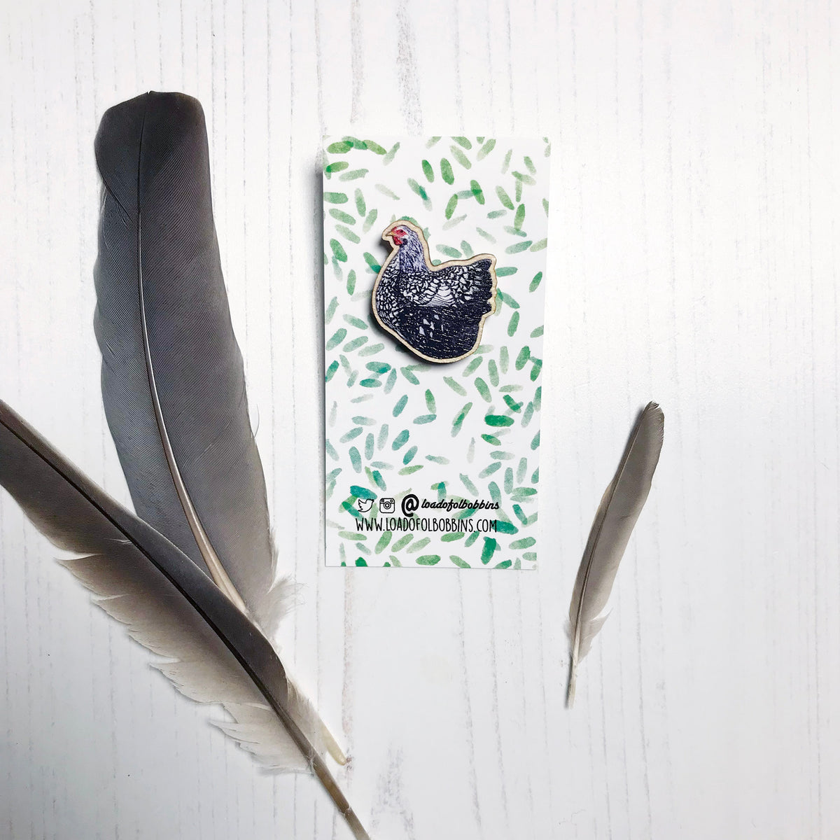 A 26mm mini wooden lapel pin of a Wyandotte chicken mounted on an illustrated backing card. The pin is made from an original illustration and is detailed and expressive in style. The bird is has a lacy black and white pattern. The backing card has a repeat green watercolour effect pattern that looks like leaves.