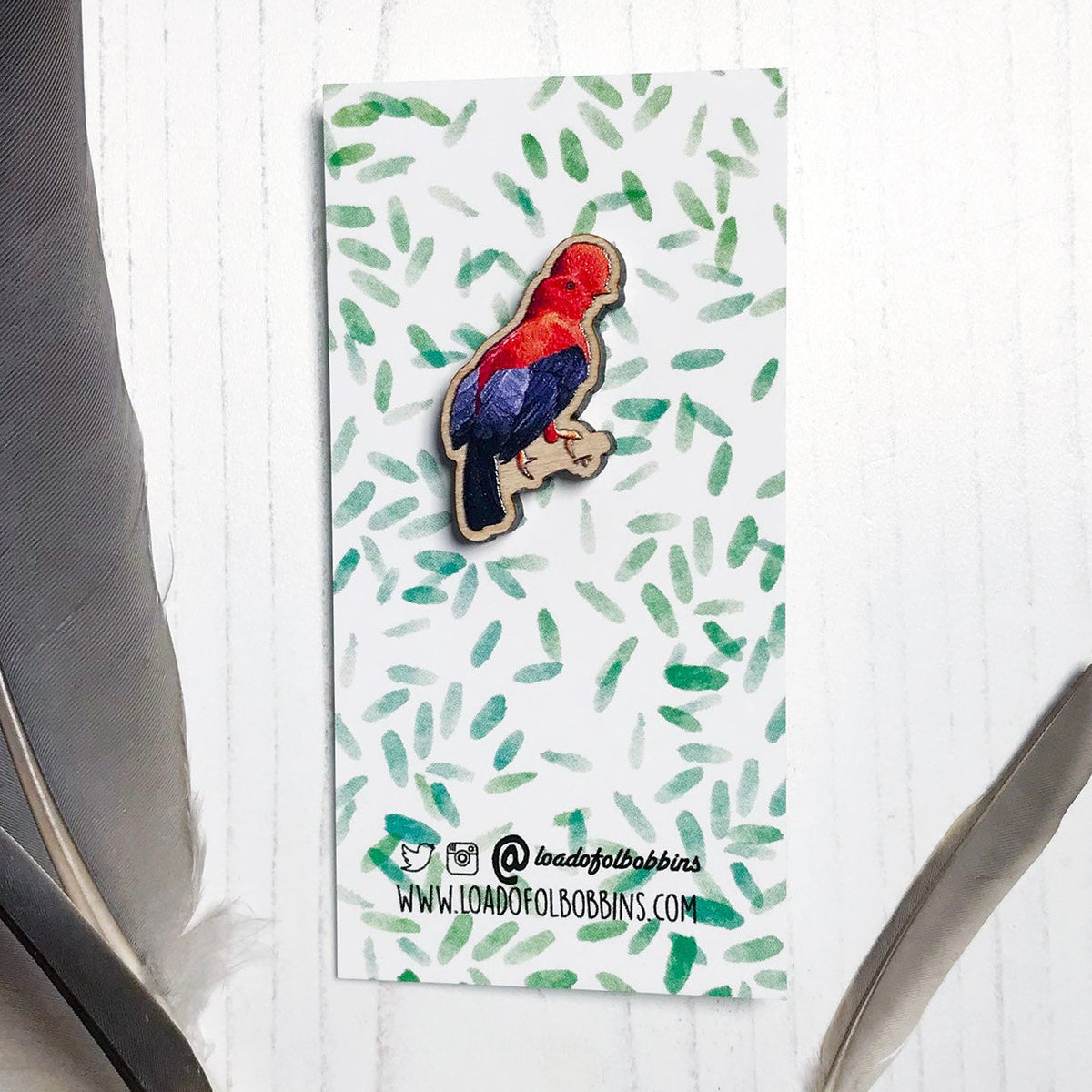 A 30mm mini wooden lapel pin of a Andean cock-of-the-rock bird perched on a branch and  mounted on an illustrated backing card. The pin is made from an original illustration and is detailed and expressive in style. The bird is orange and black with a distinct circular crest on it's head. The backing card has a repeat green watercolour effect pattern that looks like leaves.