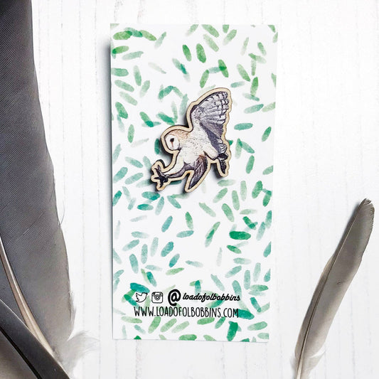 A 30mm mini wooden lapel pin of a swooping barn owl mounted on an illustrated backing card. The pin is made from an original illustration and is detailed and expressive in style. The bird is creams and browns with wings in full flight. The backing card has a repeat green watercolour effect pattern that looks like leaves.