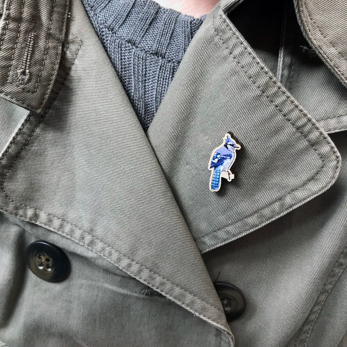 A 33mm mini wooden lapel pin of a Blue jay bird perched on a branch and  worn on the lapel of a jacket. The pin is made from an original illustration and is detailed and expressive in style. The bird is a wonderful array of blues with a small crest on it's head.