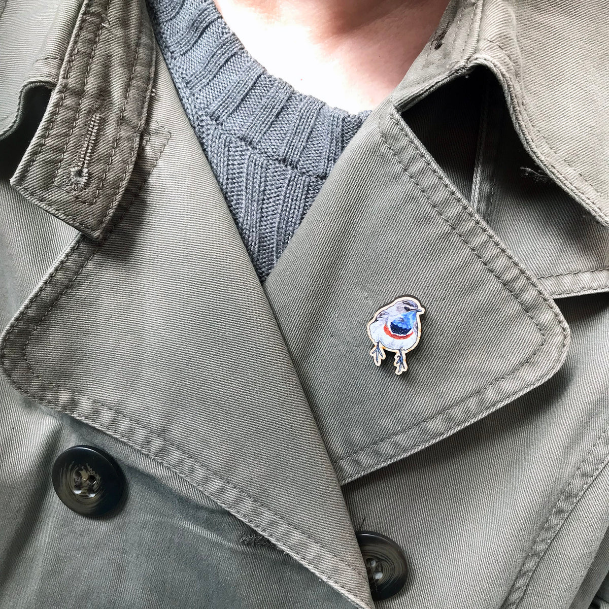 A 25mm mini wooden lapel pin of a Bluethroat bird worn on the lapel of a jacket. The pin is made from an original illustration and is detailed and expressive in style. The bird is rather robin like in appearance but with a bright blue throat.
