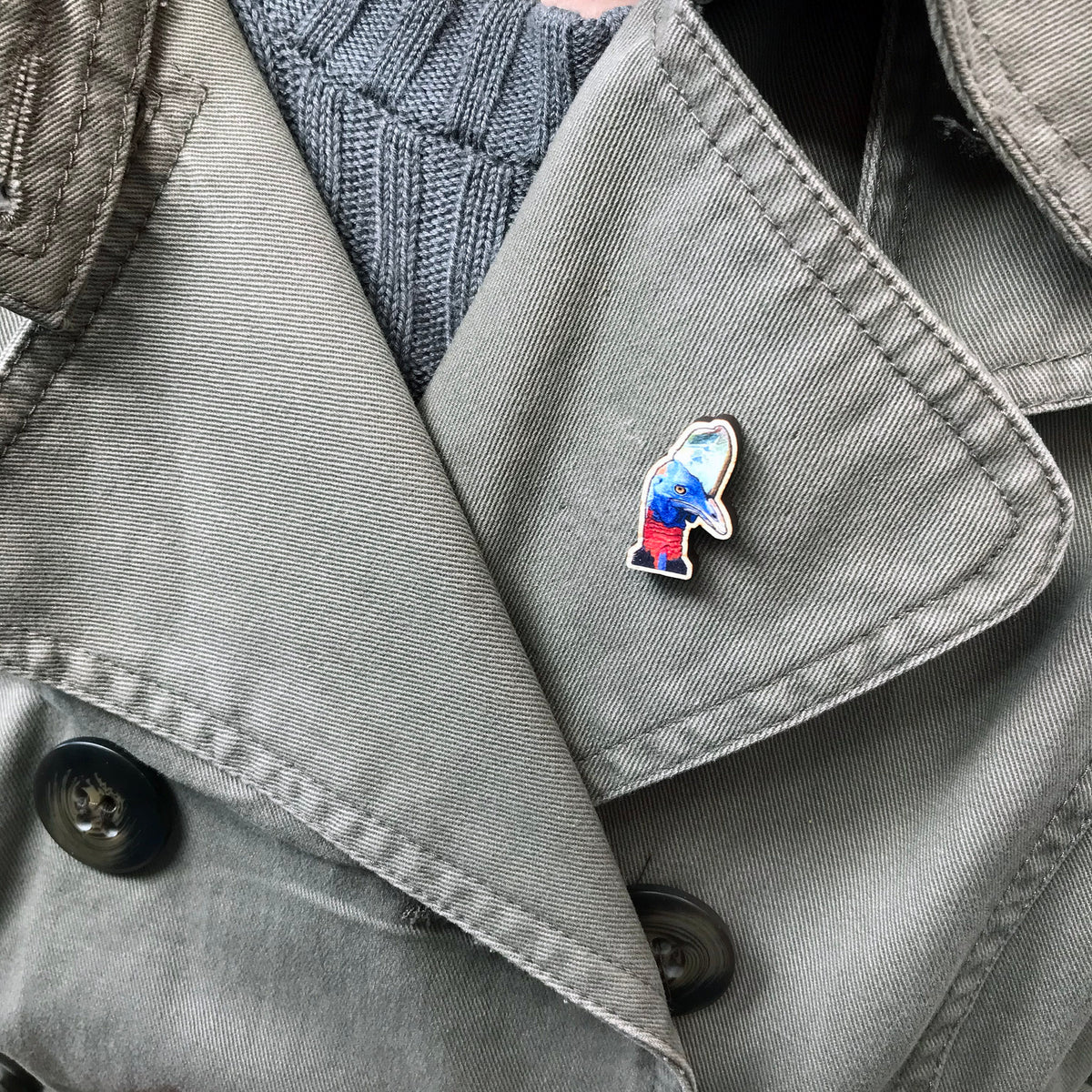 A 25mm mini wooden lapel pin of the head of a Cassowary bird worn on the lapel of a jacket. The pin is made from an original illustration and is detailed and expressive in style. The bird is rather dinosaur like in appearance with a bright blue head and red neck with blue green casque on it's head. 