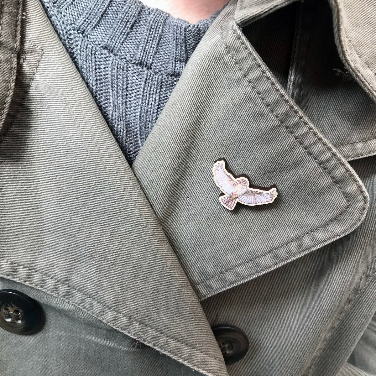 A 30mm mini wooden lapel pin of a flying female sparrow worn on the lapel of a jacket. The pin is made from an original illustration and is detailed and expressive in style. The bird is a gorgeous array of brown hues and has its wings spread wide beside it. 