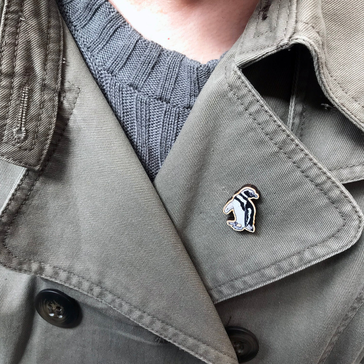 A 27mm mini wooden lapel pin of a magellanic penguin worn on the lapel of a jacket. The pin is made from an original illustration and is detailed and expressive in style. The bird has black and white markings. 