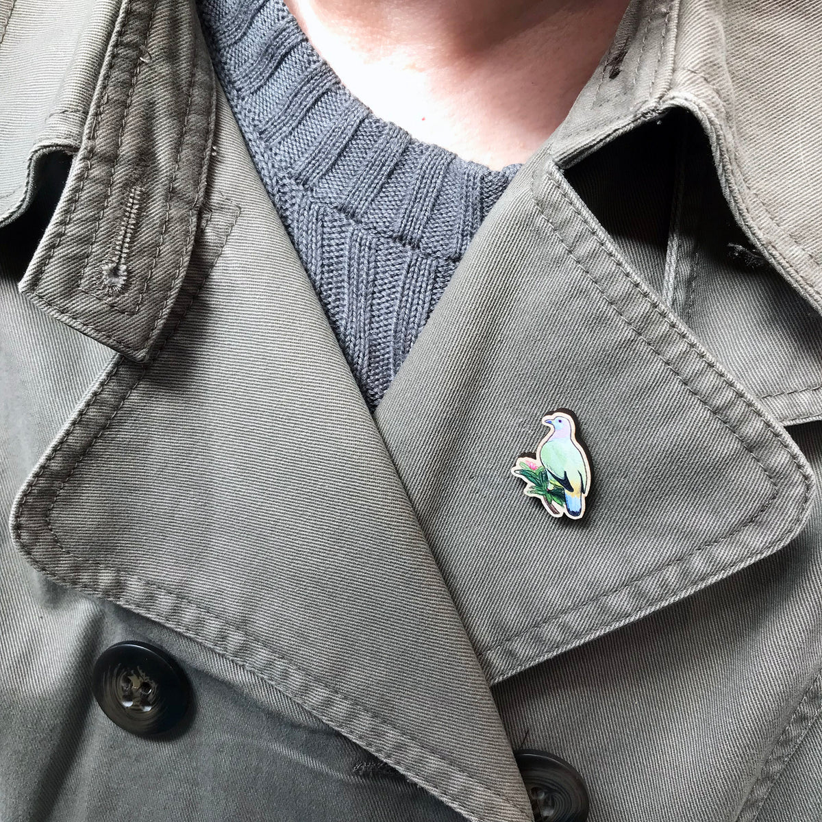 A 27mm mini wooden lapel pin of a pink-necked green pigeon worn on the lapel of a jacket. The pin is made from an original illustration and is detailed and expressive in style. The bird pastel markings in greens, pinks, yellows and blues and is perched on a lush green branch.