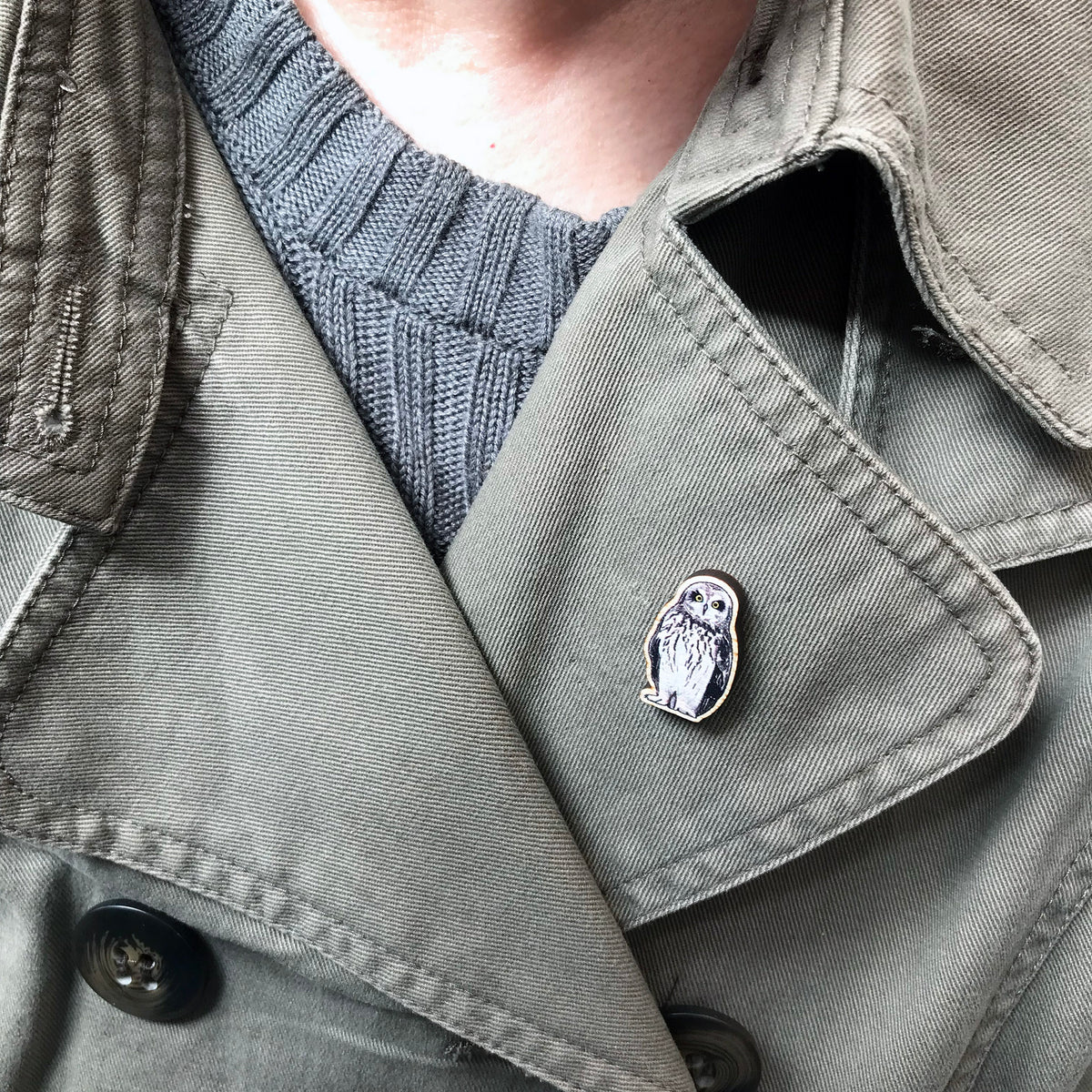 A 25mm mini wooden lapel pin of a short-eared owl worn on the lapel of a jacket. The pin is made from an original illustration and is detailed and expressive in style. The bird dark brown wings and cream front with brown flecked markings.