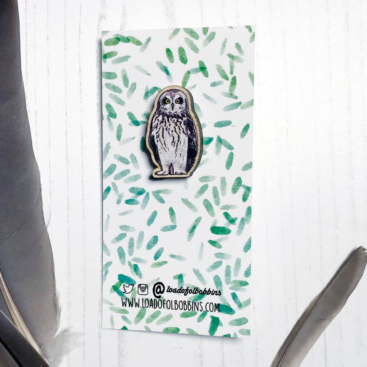 A 25mm mini wooden lapel pin of a short-eared owl mounted on an illustrated backing card. The pin is made from an original illustration and is detailed and expressive in style. The bird dark brown wings and cream front with brown flecked markings. The backing card has a repeat green watercolour effect pattern that looks like leaves.
