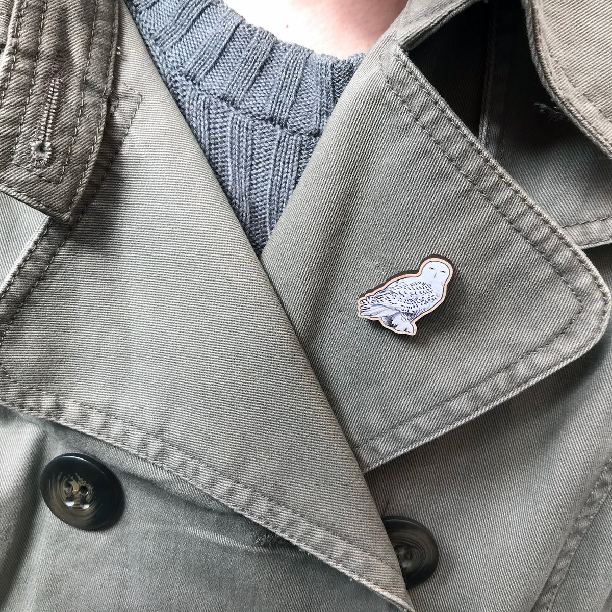 A 25mm mini wooden lapel pin of a snowy owl worn on the lapel of a jacket. The pin is made from an original illustration and is detailed and expressive in style. The bird snow white with brown flecked markings. 