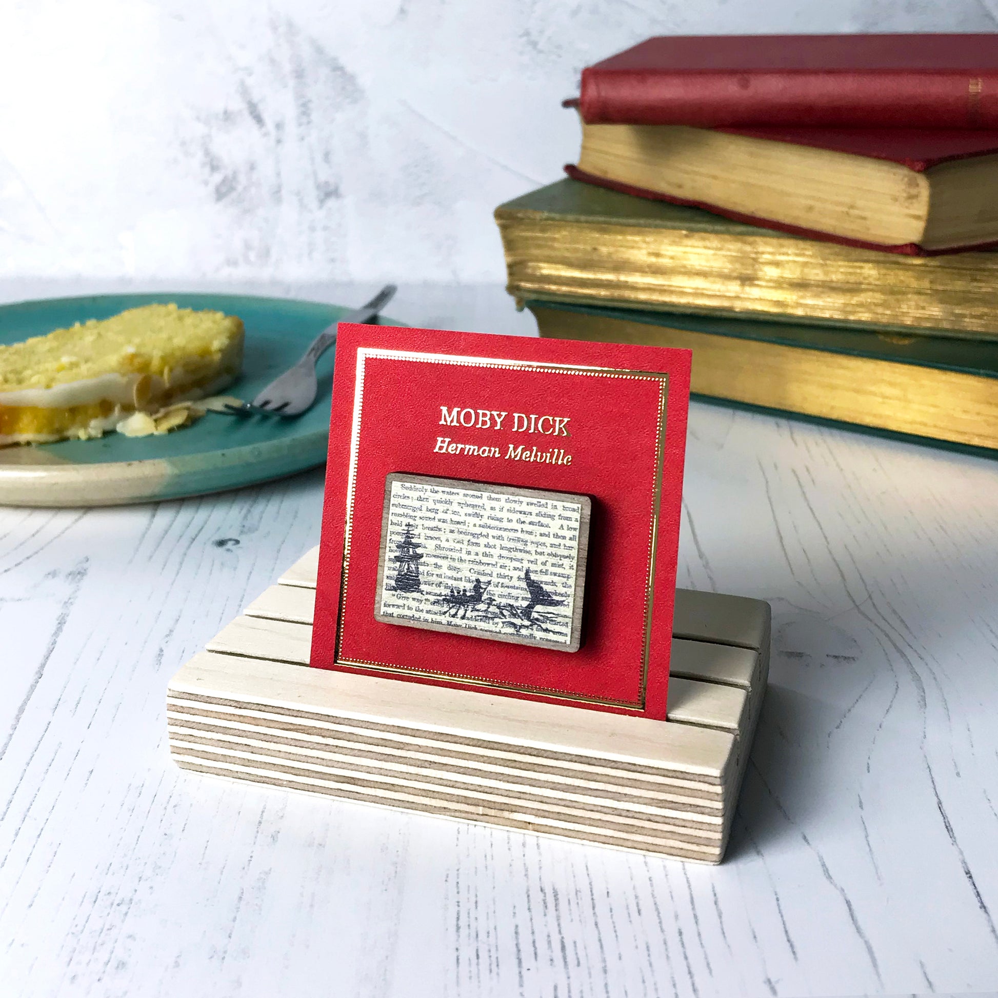 A laser cut wooden brooch showing a silhouette scene of a sailing ship and a rowing boat with men holding spears in it chasing after a large whale tail as it dives below the sea. It is inspired by the novel Moby Dick by Herman Melville and is printed with the text from the novel behind it. The necklace is mounted on a red backing card with gold foil lettering detail and made to look like the front of a book.