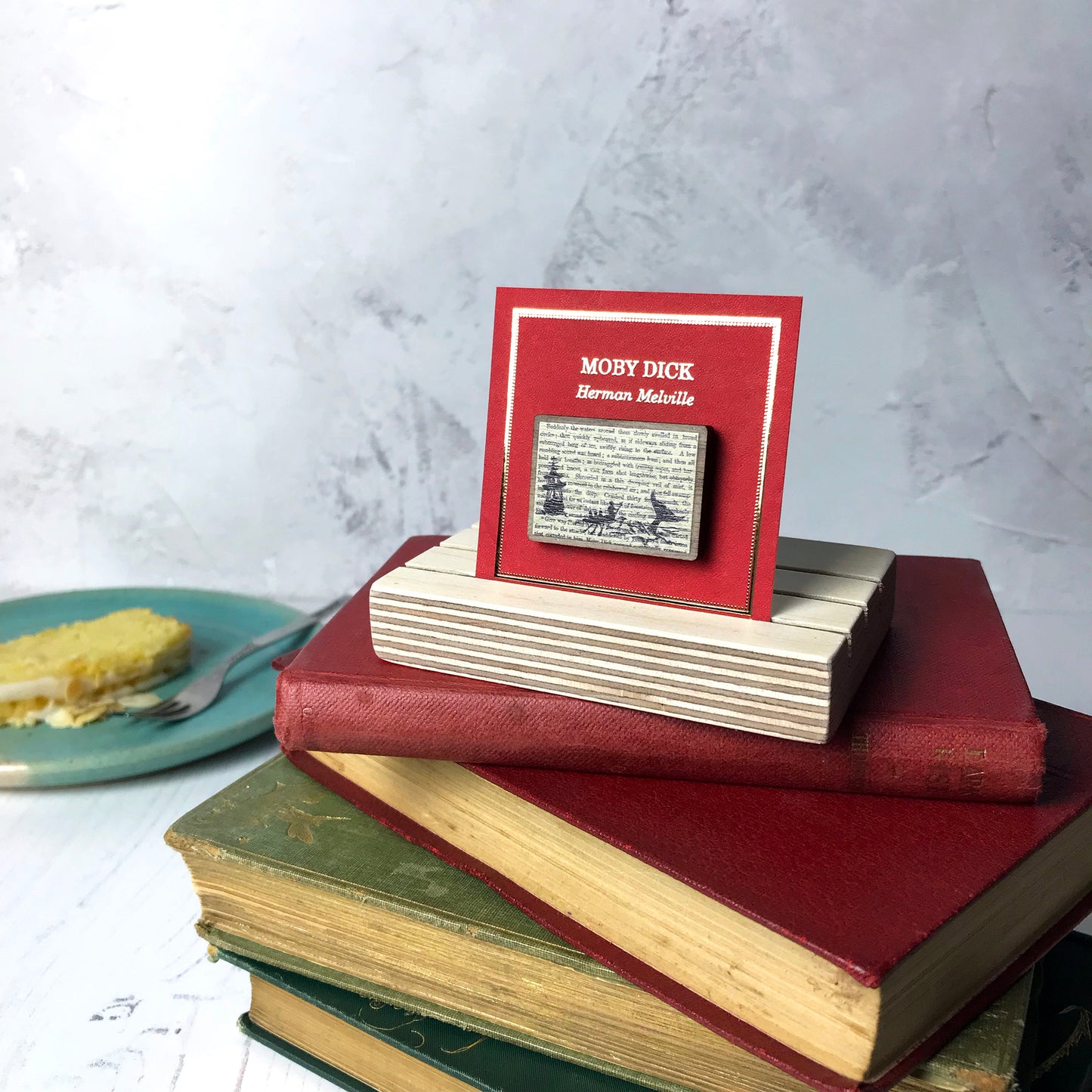 A laser cut wooden brooch showing a silhouette scene of a sailing ship and a rowing boat with men holding spears in it chasing after a large whale tail as it dives below the sea. It is inspired by the novel Moby Dick by Herman Melville and is printed with the text from the novel behind it. The necklace is mounted on a red backing card with gold foil lettering detail and made to look like the front of a book.
