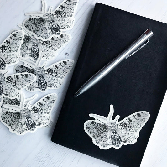 A peppered moth sticker created from an original digital illustration by Loadofolbobbins. A fairly large moth which has mottled black markings against white allover it's wings, body and legs. It has light colour, feather looking, antennae. The whole moth looks a little bit like a rorschach test . The moth has it's wings out, frozen in a moment of flapping. The sticker is on a black notebook with a pen resting on it.