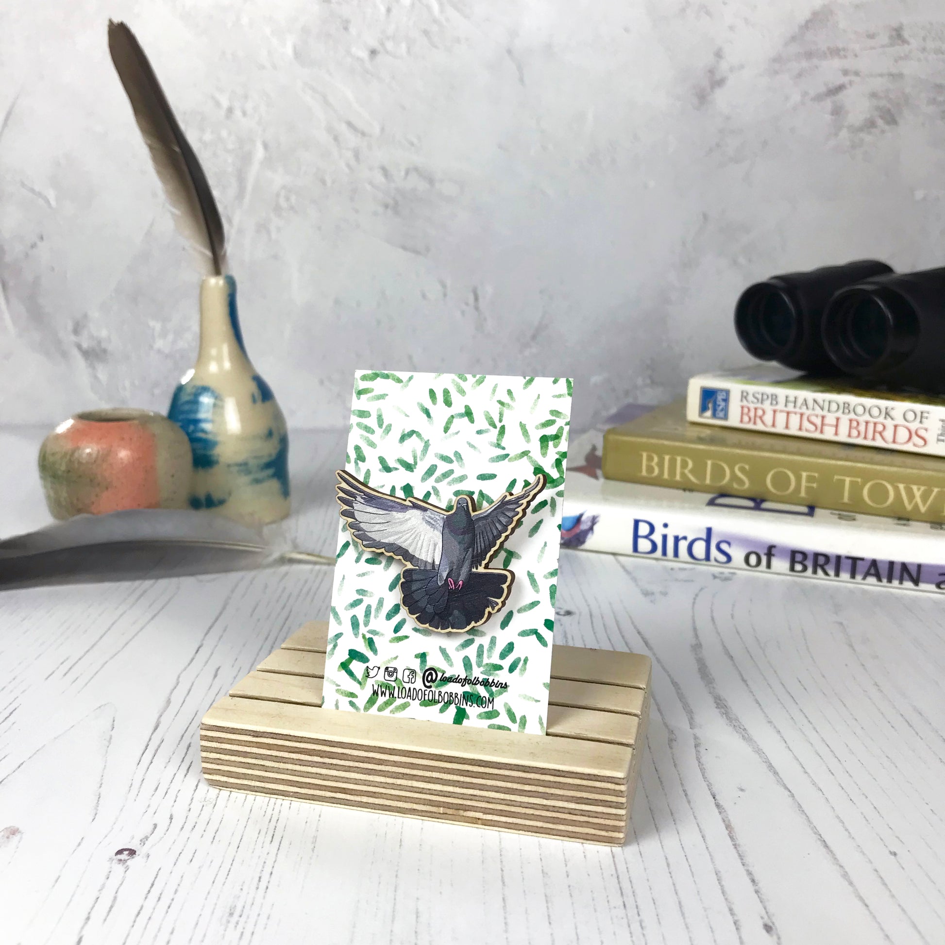 A laser cut wooden brooch showing an illustration of a flying pigeon bird by loadofolbobbins. The brooch is mounted on a backing card with a repeat abstract watercolour leaf pattern. They are standing in a wooden stand and in the background you can see a pile of bird watching books and a pair of binoculars.
