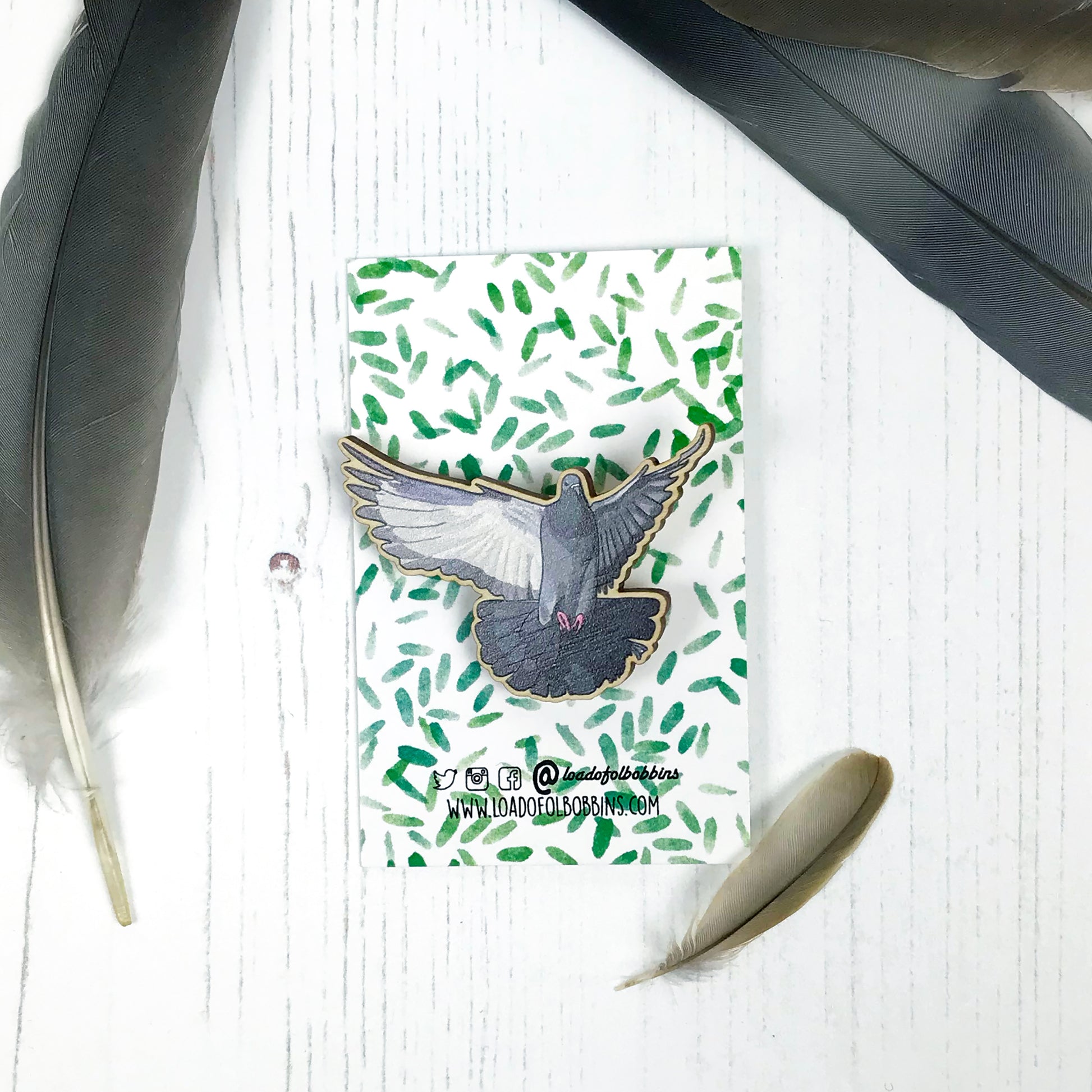 A laser cut wooden brooch showing an illustration by Loadofolbobbins of a flying pigeon with wings spread out wide. The brooch is mounted on a backing card with a repeat abstract watercolour leaf pattern.