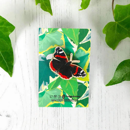 A laser cut wooden brooch showing an illustration by Loadofolbobbins of a red admiral butterfly. The brooch is mounted on a backing card with a repeat ivy leaf pattern.